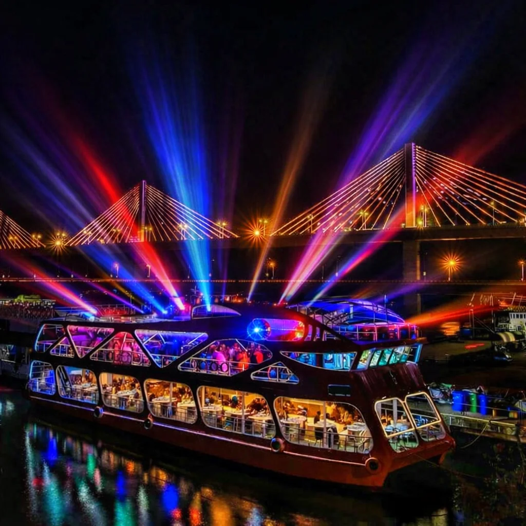 Dinner cruise boat glowing with coloured spotlights at night, guests dining inside and dancing on deck