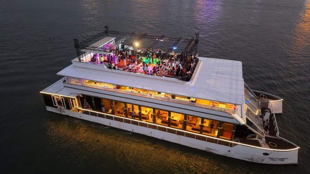 Guests dancing on the open-air upper deck of a lit cruise boat on the Mandovi river at night